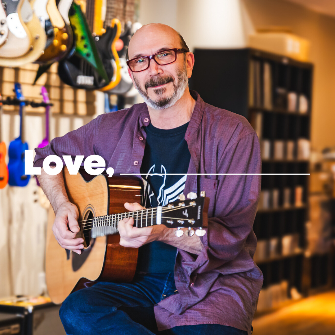 A smiling man with brown hair and a beard, dressed in a black t-shirt with a purple shirt over it and jeans, holds a guitar and sits on a stool in a shop with guitars hanging on the wall behind him.